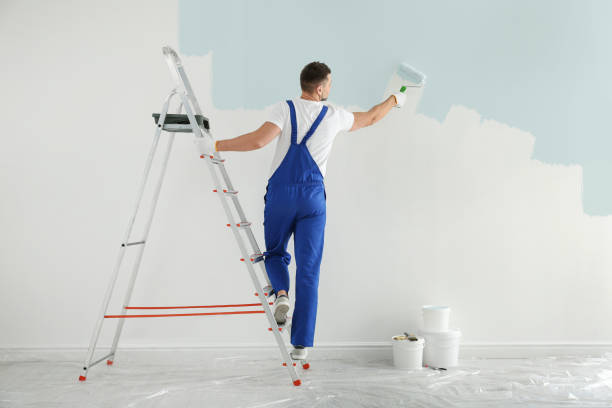 A worker in blue overalls paints a pale blue wall from a step ladder, with paint buckets and protective sheeting below; bright indoor lighting.