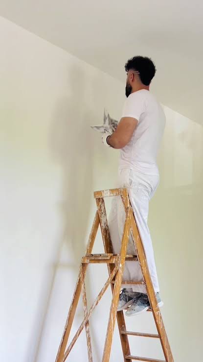 A worker in white clothing stands on a wooden step ladder and applies material to an indoor wall; soft, even lighting.